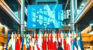 Werden Europäer zu Kulturbanausen? Europäer als Kulturbanausen? Ein Mann in kurzen Hosen und Rücksack schreitet vorbei an einer reihe von Fahnen mit der EU-Fahne im Hintergrund