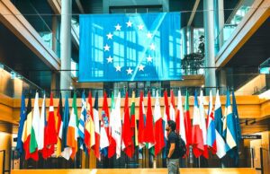 Werden Europäer zu Kulturbanausen? Europäer als Kulturbanausen? Ein Mann in kurzen Hosen und Rücksack schreitet vorbei an einer reihe von Fahnen mit der EU-Fahne im Hintergrund