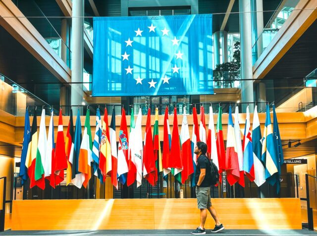 Europäer als Kulturbanausen? Ein Mann in kurzen Hosen und Rücksack schreitet vorbei an einer reihe von Fahnen mit der EU-Fahne im Hintergrund