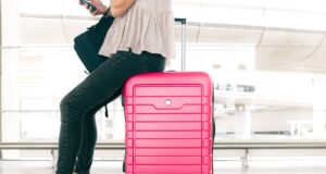 Flugüberbuchung: Recht auf Schadenersatz Frau in langer schwarzer Hose und mit Bluse sitzt auf einem pinken Schalenkoffer am Flughafen