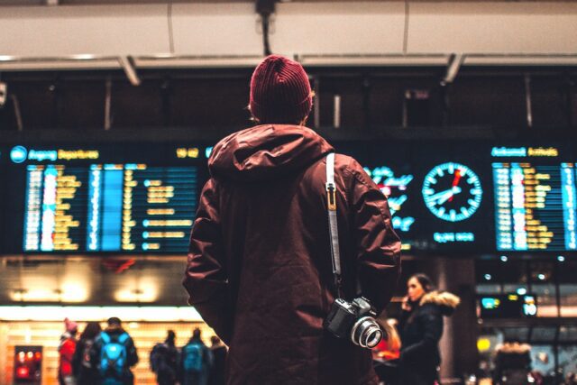 Travellyze-Studie: Reisebranche im Aufwind: Bild zeigt Mann am Flughafen mit Kamera vor Abfluganzeige