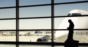 Wartezeiten auf Dienstreisen clever nutzen Wartezeiten clever überbrücken: Bildzeigt einen Geschäftsreisenden am Flughafen mit Blick auf ein großes Flugzeug auf dem Flugfeld