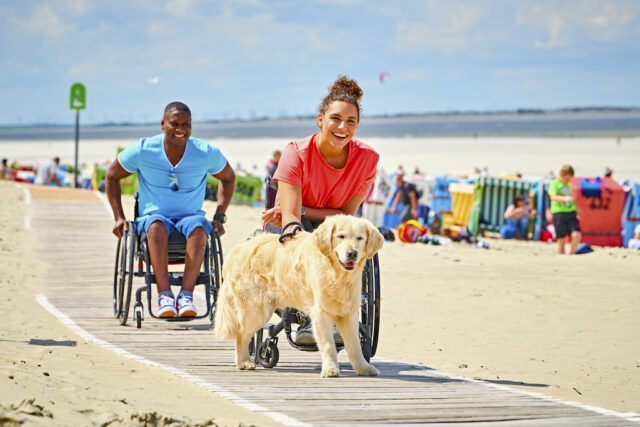 Barrierefrei reisen: Bild zeigt einen Mann und eine Frau im Rollstuhl mit einem Hund am Strand von Langeoog