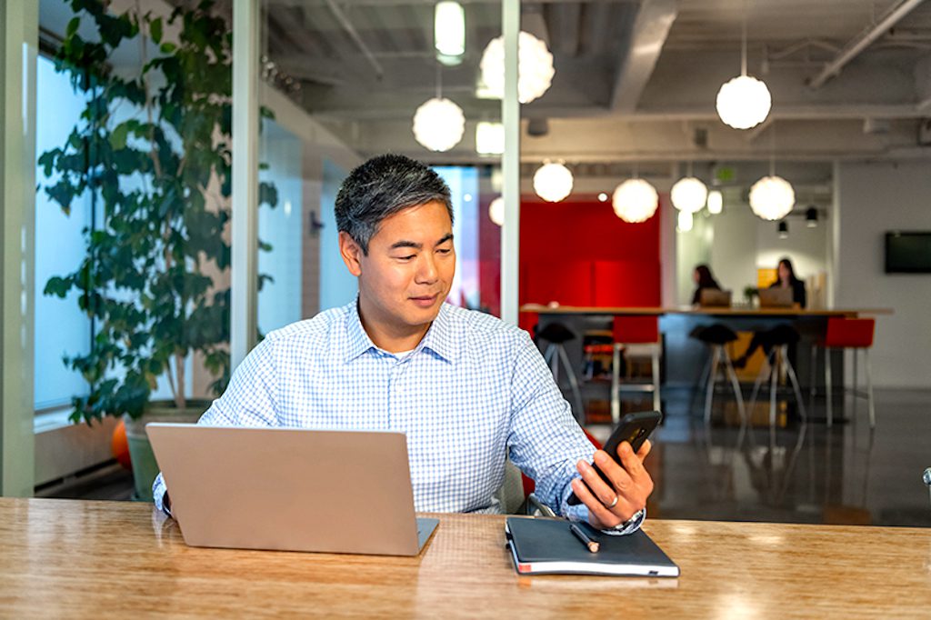 Travel Manager werden von Unternehmen unterschätzt: Bild zeigt ein Office mit einem Mann vor seinem Laptop und mit einem Smartphone in der Hand