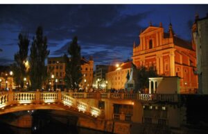 In Ljubljana ein neues Hotelgefühl erleben Ljubljana bei Nacht. Beleuchtete Brücke über den Fluss Ljujlanica, Abendstimmung, festlich.