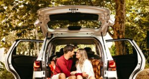Checkliste für eine sichere Urlaubsfahrt Ein junger Mann und eine junge Frau sitzen im Gepäckraum ihres Autos mit geöffneten die Türen
