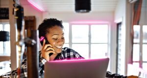 Business-Trends: AI und Smart Office gefragt Business-Trends: Dunkelhäutige Frau sitzt vorm Bildschirm und telefoniert. Sie lacht dabei