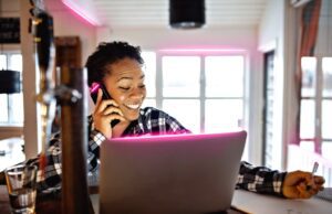 Business-Trends: AI und Smart Office gefragt Business-Trends: Dunkelhäutige Frau sitzt vorm Bildschirm und telefoniert. Sie lacht dabei