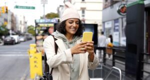Reise-App führt zu den schönsten Plätzen Mobile Reise-App für Städtebummler: Junge Frau mit Mütze, gelbem Rucksack und gelbem Smartphone lässt sich von der App zum Ziele führen