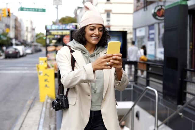 Mobile Reise-App für Städtebummler: Junge Frau mit Mütze, gelbem Rucksack und gelbem Smartphone lässt sich von der App zum Ziele führen