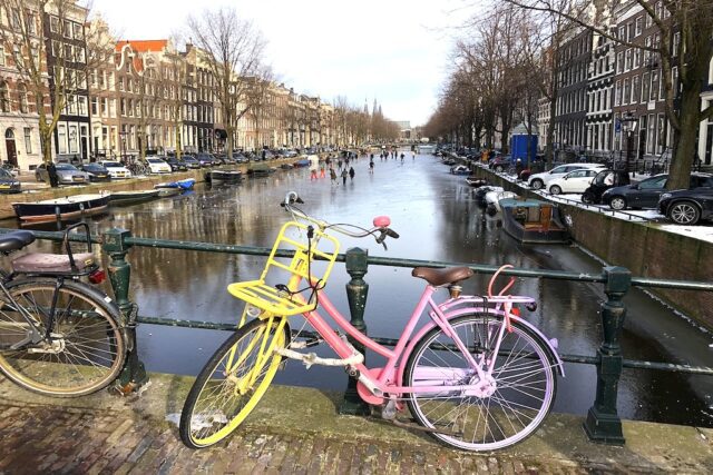 Nach Amsterdam mit Eurowings ab Salzburg jetten. Zusehen ist eine vereist Gracht. Am Brückengeländer geht ein buntes Fahrrad. Auf der vereisten Gracht laufen Menschen Schlittschuh