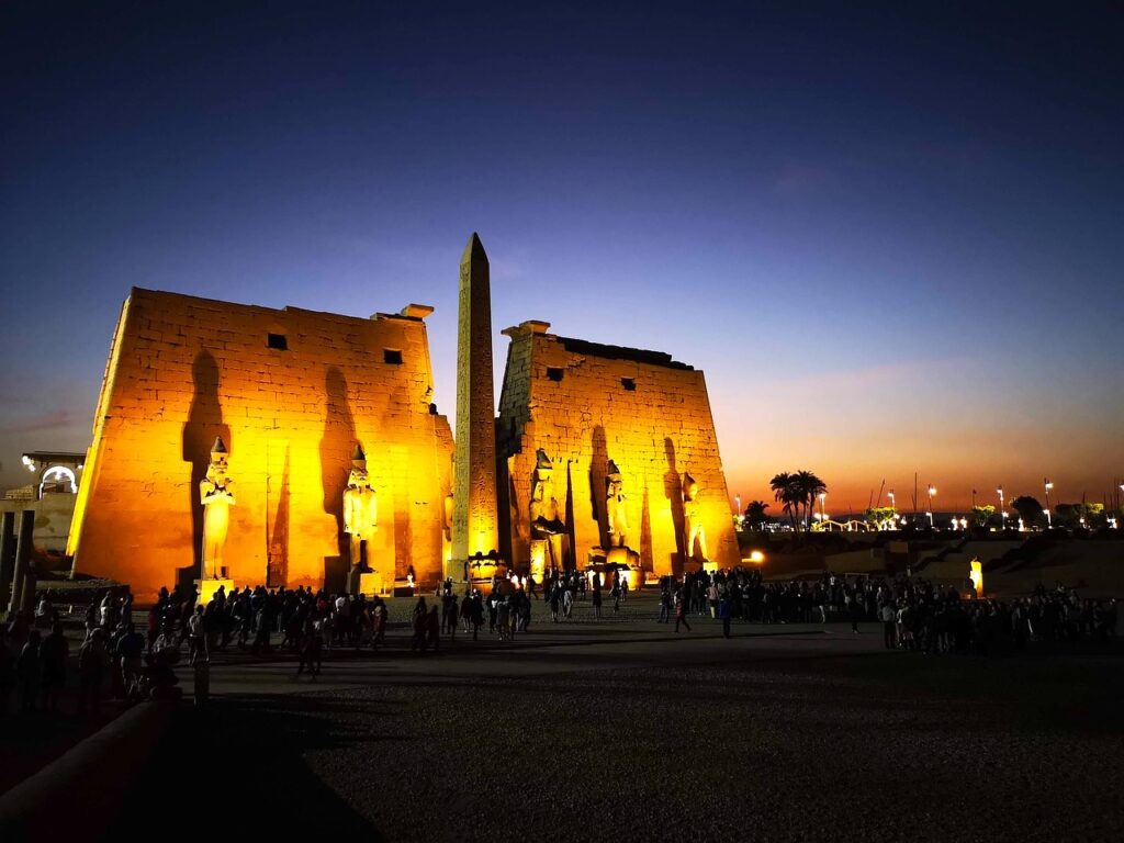 Der berühmte Tempel von Luxor steht auf dem Programm jeder Nil-Kreuzfahrt. Zu sehen ist der festlich beleuchtete Tempel fünf hohen Figuren im Vordergrund  in der Abenddämmerung
