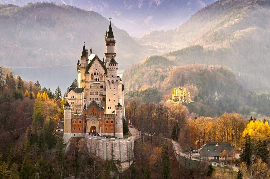 Im Bild ist das legendäre Schluss Neuschwanstein in einer herbstlichen Landschaft aus Wäldern und Bergen zu sehen