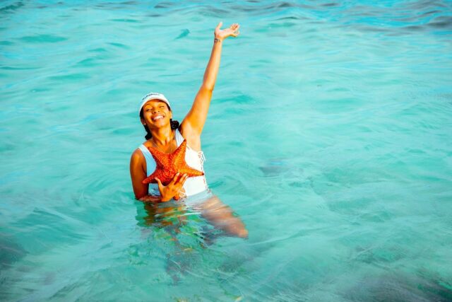 Frühbucher profitieren bei den Reisekosten: Frau im türkisfarbenen Meer streckt eine Hand zum Himmelt und mit der anderen hält sie einen roten Seestern vor dem Badekostüm