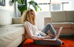 Legalisierung des Online Glücksspiels 2024 Legalisierung des Online Glücksspiels: Junge Frau sitzt – angelehnt an einer Couch - auf einem roten Teppichboden und tippt auf ihren Laptop