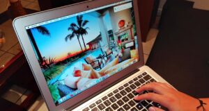 Betrügereien auf Buchungsplattformen Betrügereien auf Buchungsplattformen: Laptop mit einem Hotelbild in der Karibik auf dem Bildschirm, zwei Hände tippen auf der schwarzen Tastatur