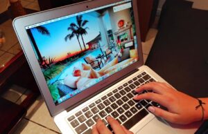 Betrügereien auf Buchungsplattformen Betrügereien auf Buchungsplattformen: Laptop mit einem Hotelbild in der Karibik auf dem Bildschirm, zwei Hände tippen auf der schwarzen Tastatur