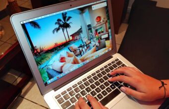 Neue Betrugsmaschen bei Reisebuchungen Betrugsmaschen auf Buchungsplattformen: Laptop mit einem Hotelbild in der Karibik auf dem Bildschirm, zwei Hände tippen auf der schwarzen Tastatur