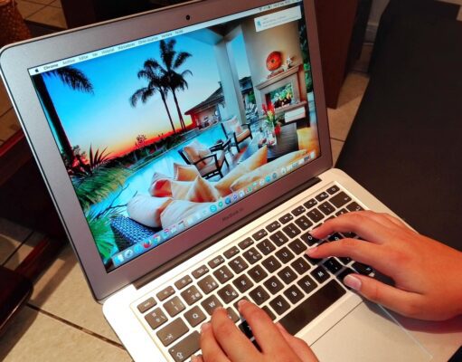Neue Betrugsmaschen bei Reisebuchungen Betrugsmaschen auf Buchungsplattformen: Laptop mit einem Hotelbild in der Karibik auf dem Bildschirm, zwei Hände tippen auf der schwarzen Tastatur