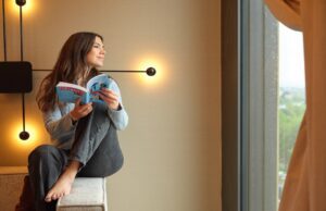 Adina Hotels launcht Mitgliederprogramm Adina Hotels verbessert das Mitgliederprogramm: Kunde Frau sitzt mit einem Buch auf einer Couch-Armlehne und blickt im von einer Wandleuchte beleuchteten Hotelzimmer in Richtung Fenster