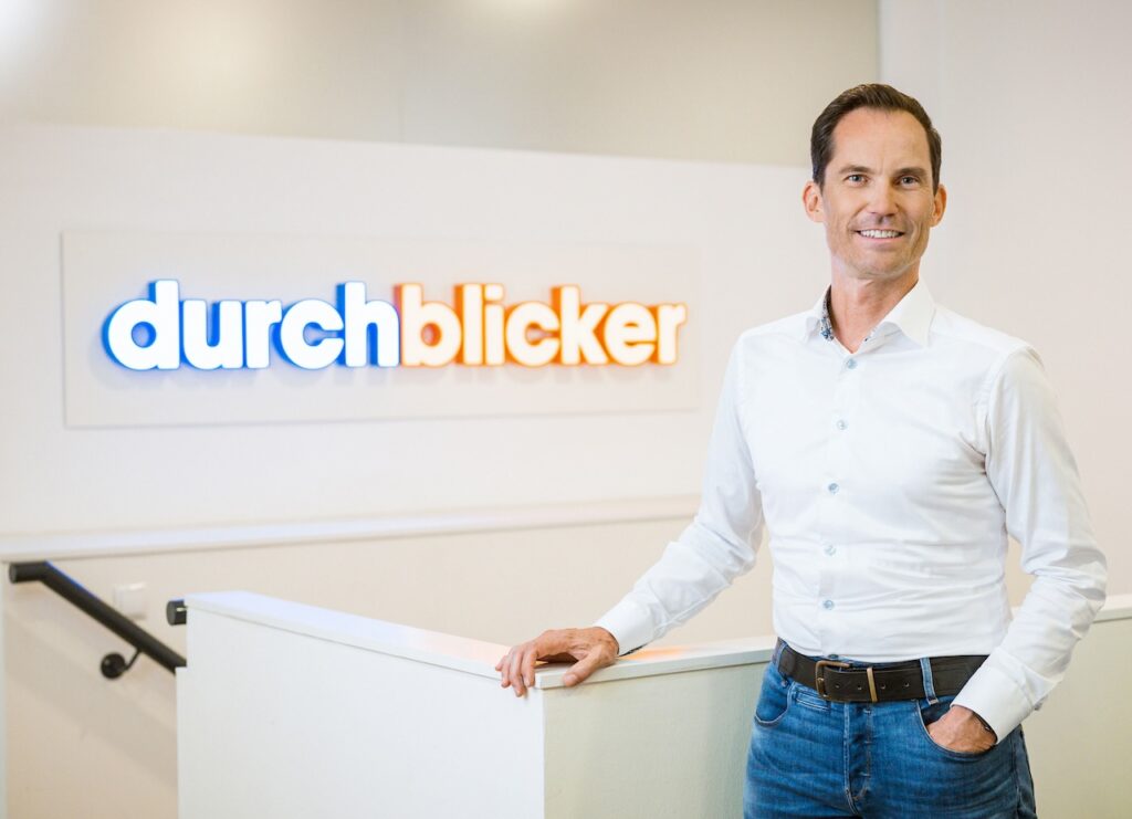 Bild zeigt Martin Spona stehend vor einer Wand mit dem Firmenlogo „durchblicker“