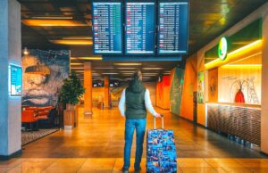 Urlaubsbudget heuer um 300 Euro geringer Mageres Urlaubsbudhet: Reisender in Jeans mit Koffer steht in der Abflughalle und blickt auf eine Anzeigetafel mit den Abflugzeiten der Airlines