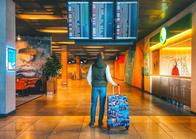 Mageres Urlaubsbudhet: Reisender in Jeans mit Koffer steht in der Abflughalle und blickt auf eine Anzeigetafel mit den Abflugzeiten der Airlines
