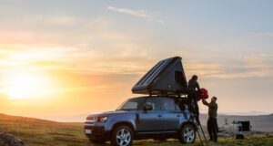 Mit dem Defender dem Alltag entfliehen Sonnenuntergang. Das Dachzelt auf dem Dach des Defender ist hochgeklappt, zwei Personen stehen am Heck des Autos.