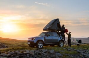 Mit dem Defender dem Alltag entfliehen Sonnenuntergang. Das Dachzelt auf dem Dach des Defender ist hochgeklappt, zwei Personen stehen am Heck des Autos.