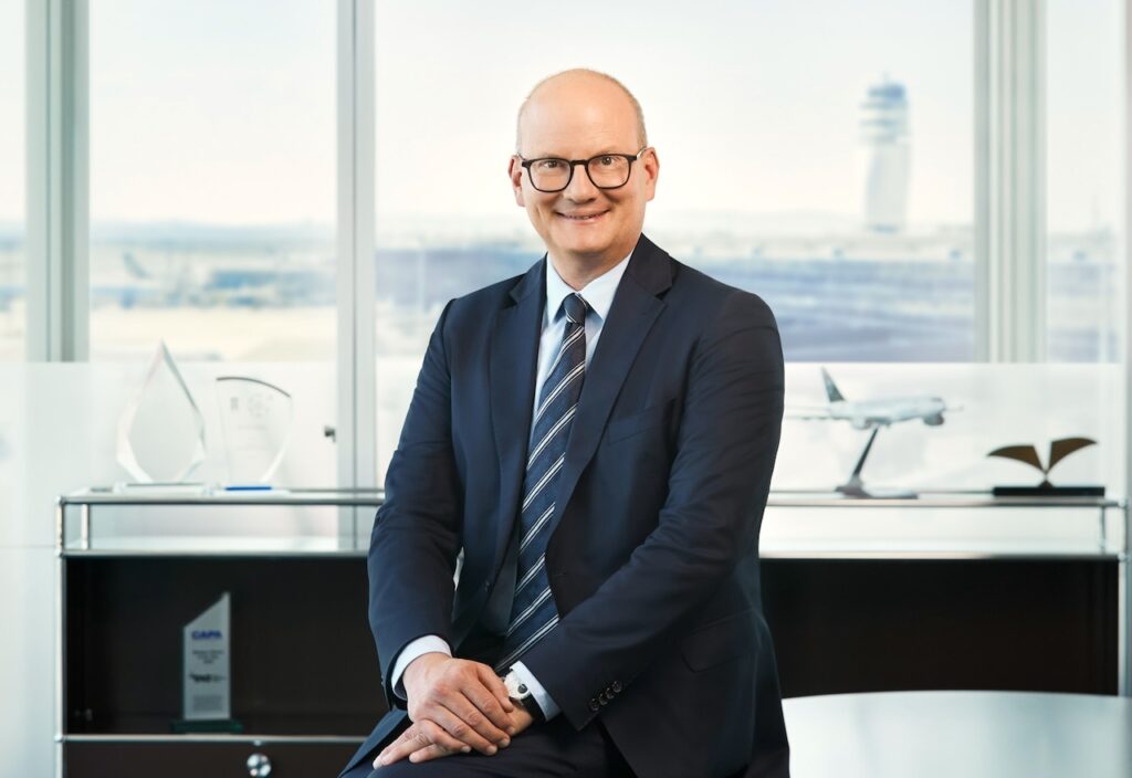 Magister Julian Jäger sitzt an einem Tisch mit gekreuzten Händen, im Hintergrund ist der Flughafen Wien zum sehen