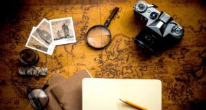 Urlaubsplanung und Künstliche Intelligenz Historische Weltkarte, darauf Kamera, Fotos, Sonnebrillem, Lupe, Fotos und Notizbuch mit Bleistift