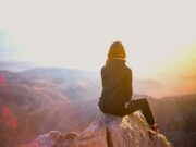 Bewusst reisen beginnt mit Bewusstsein Bewusst reisen beginnt mit Bewusstsein: Eine Frau sitzt auf einem einem Felsen eines Gipfels. Sie blickt in eine Berglandschaft im Licht der untergehenden Sonne: Nachhaltig reisen ist in