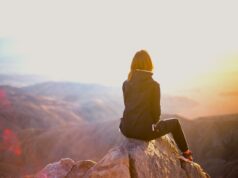 Bewusst reisen beginnt mit Bewusstsein Bewusst reisen beginnt mit Bewusstsein: Eine Frau sitzt auf einem einem Felsen eines Gipfels. Sie blickt in eine Berglandschaft im Licht der untergehenden Sonne: Nachhaltig reisen ist in