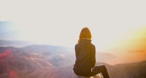 Nachhaltig reisen ist eine Sache des Wollens Eine Frau sitzt auf einem einem Felsen eines Gipfels. Sie blickt in eine Berglandschaft im Licht der untergehenden Sonne: Nachhaltig reisen ist in