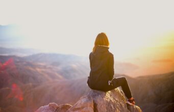 Bewusst reisen beginnt mit Bewusstsein Bewusst reisen beginnt mit Bewusstsein: Eine Frau sitzt auf einem einem Felsen eines Gipfels. Sie blickt in eine Berglandschaft im Licht der untergehenden Sonne: Nachhaltig reisen ist in