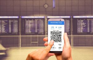 Zahlen Konsumenten für ihre Flüge zuviel? Mann hält ein Handy und tippt mit Zeigefinger das digitale Flugticket auf dem Display. Im Hintergrund ist eine riesige Anzeigetafel mit Ankünften und Abflügen am Flughafen zu sehen