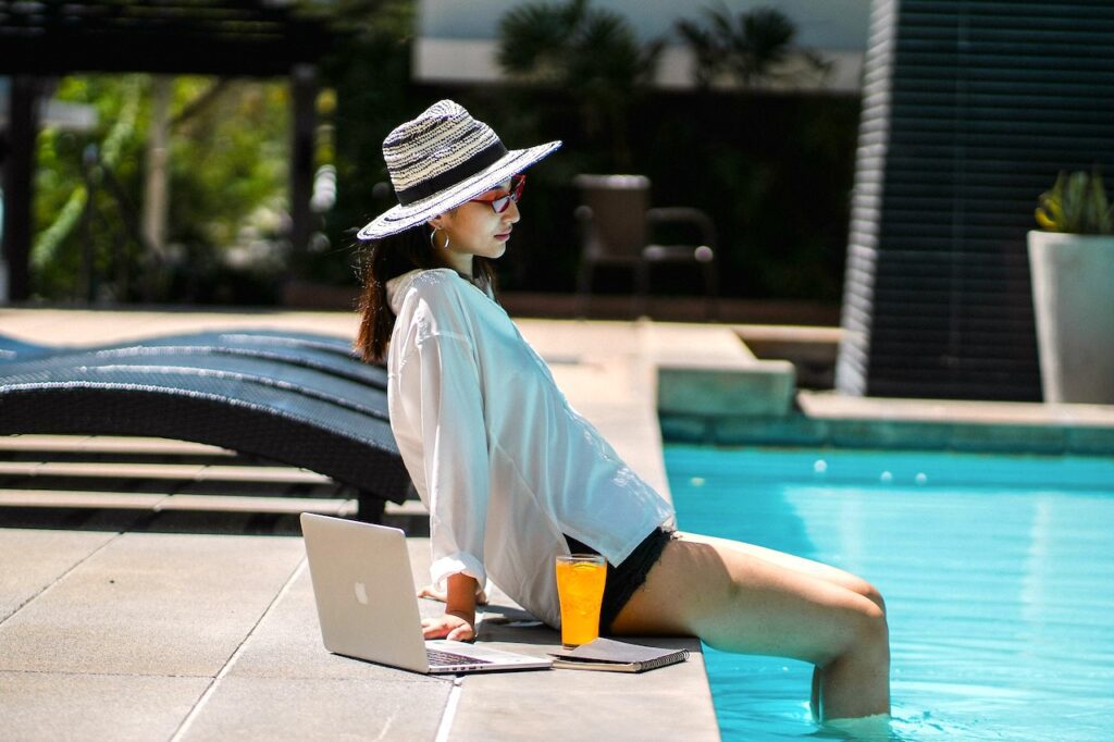 junge Frau mit Sonnenbrille und Hut sitzt neben ihrem Laptop und einem Glas mit Orangensaft am Rand eines Pools mit türkisen Wasser gefüllten Beckens