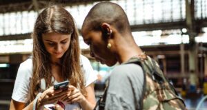 Die digitale Zukunft des Reisens ist schon da! Zwei junge Frauen mit Riucksack schauen auf ihre Mobiltelefone in einer Abflughalle und suchen nach Informationen - so sieht bereits heute die digitale Zukunft des Reisens aus