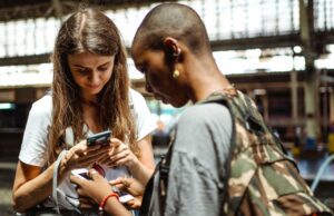 Die digitale Zukunft des Reisens ist schon da! Zwei junge Frauen mit Riucksack schauen auf ihre Mobiltelefone in einer Abflughalle und suchen nach Informationen - so sieht bereits heute die digitale Zukunft des Reisens aus