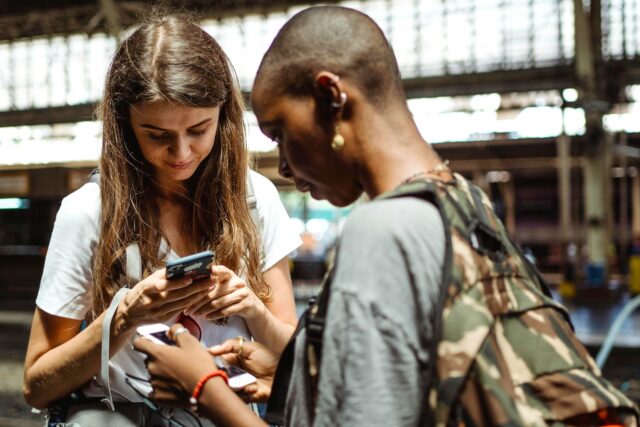 Digitale Zukunft des Reisens Zwei junge Frauen mit Riucksack schauen auf ihre Mobiltelefone in einer Abflughalle und suchen nach Informationen - so sieht bereits heute die digitale Zukunft des Reisens aus