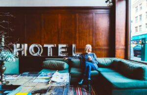 Das Hotel der Zukunft mit vielen Aussichten Frau sitzt auf einer Türkisen Couch in einer holzgetäfelten Hotellobby und blickt beim Fenster auf die Straße, hinter ihr Buchstaben eines alten Neonschildes mit den Buchstaben für Hotel