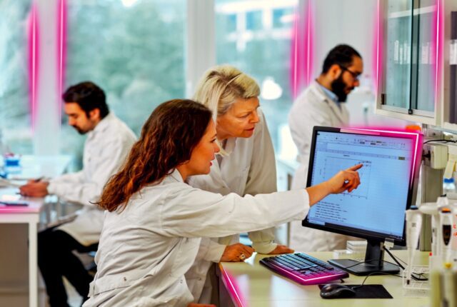 MenschenIn einem Büro, wo eine Frau vor einem Computer sitzt und einer Kollegin etwas auf dem Bildschirm zeigt