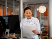 KI-Kompetenz fehlt bei Geschäftsreisen KI-Kompetenz:Asiatisch aussehende Businessfrau im weißen Kostüm steht vor einem Computerbildschirm und blickt auf ein Blatt Papier