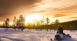 In Lappland lockt noch das wahre Abenteuer Winterlandschaft in Lappland, wolkenbehangener Himmel durch den die Sonne scheint und ein Schneemobil auf einer Schneepiste