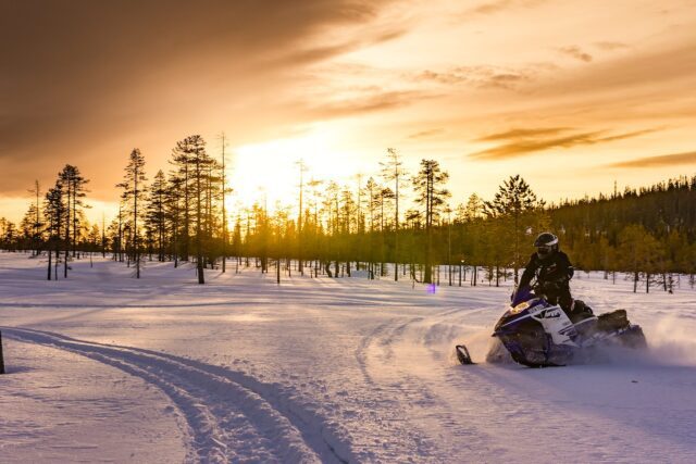 Winterlandschaft in Lappland, wolkenbehangener Himmel durch den die Sonne scheint und ein Schneemobil auf einer Schneepiste