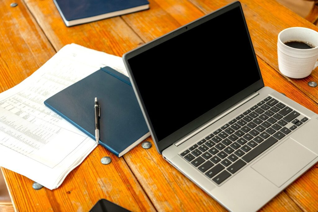Ein Laptop mit schwarten Bildschirm, daneben ein dunkelgrüner Notizblick mit einem Stift, diverse Unterlagen und eine volle Kaffeetasse