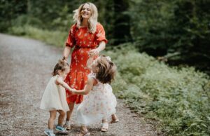 Wenn Mütter über Urlaubsreise entscheiden Mütter entscheiden über Urlaubsziele: Eine Frau in einem rotgetupften Kleid spielt mit zwei kleinen Mädchen in weißen Kleidern auf einem Waldweg