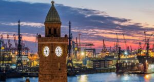 Hamburg: Viel Chic, Charme und Tradition Bild zeigt den Hamburger Hafen in der Dämmerstunden mit dunkelblauem und rotem Himmel. Im Vordergrund die Landungsbrücken und im Hintergrund Docks, Schiffe, und Kräne