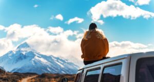 Soloreisen mit dem Camper liegt im Trend Soloreisen mit dem Camper: Frau in einem gelben Anorak sitzt auf dem Dach eines Campers und betrachtet schneebedeckte Gipfel' in der Ferne