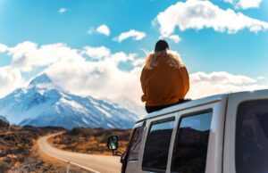 Soloreisen mit dem Camper liegt im Trend Soloreisen mit dem Camper: Frau in einem gelben Anorak sitzt auf dem Dach eines Campers und betrachtet schneebedeckte Gipfel' in der Ferne